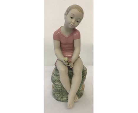 A large Nao ceramic figurine of a girl sitting on a rock, wearing a pink dress and holding flowers.  In matt and gloss finish