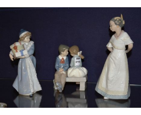 Three Nao figures, in the form of a child carrying presents, 23cm high, boy and girl in embrace sitting on bench, 17cm high a