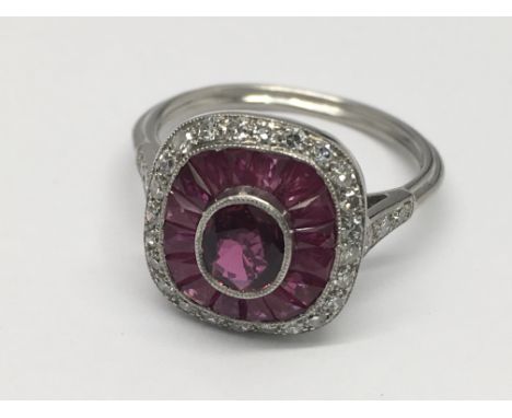 A platinum Art Deco style ruby and diamond ring. Central oval-cut ruby surrounded by a Hal of calibre cut rubies with a filig