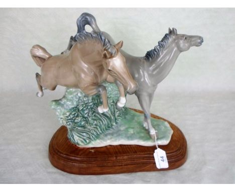 A Nao figure group of two horses jumping a hedge, mounted to bespoke wooden stand, 29cm tall.