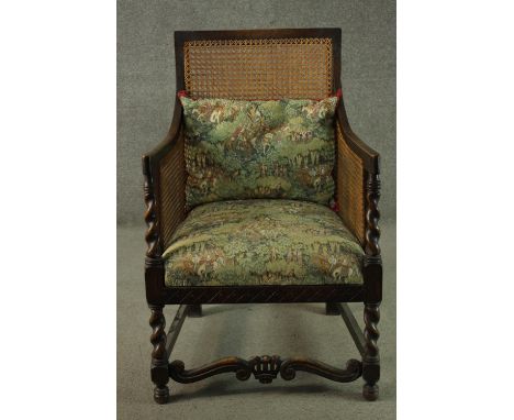 An early 20th century oak bergere armchair, with a caned back and arms, the seat and loose cushion upholstered in a tapestry 