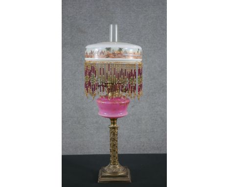 A 19th century brass oil lamp, with an ornate brass column base, opaque pink glass font and an Egyptian design beaded fringed