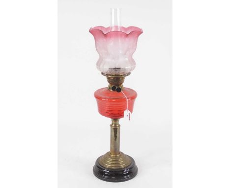 An early 20th century oil lamp, having an associated cranberry tinted etched glass shade above a red glass font, on brass ped