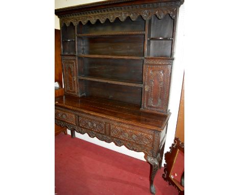 A 17th century style low relief carved oak dresser, having a wavy floral carved frieze over open shelving to twin arched pane