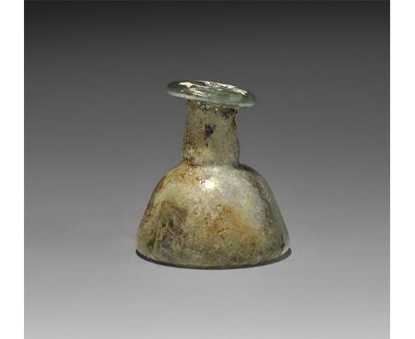  1st-3rd century AD. A squat iridescent glass bell-shaped body with concave base, short neck, eccentric folded rim. Cf. White