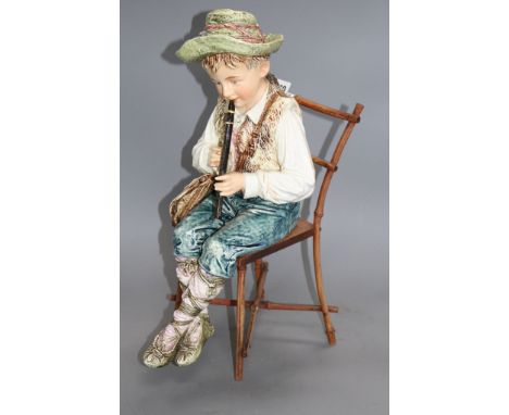 A late 19th century Austrian tin glaze pottery figure of a Tyrolean boy playing a flute, seated upon a bamboo wood chair, hei