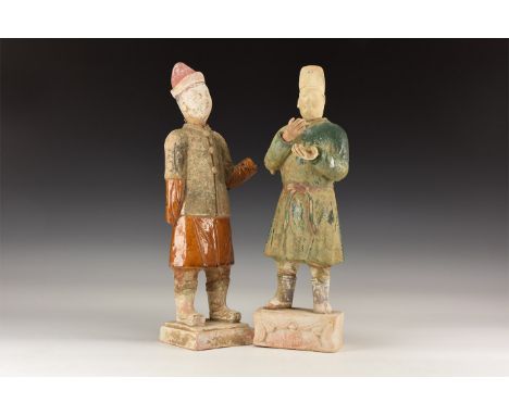 An undated archaistic pair of ceramic statuettes of male attendants wearing caps, tunics and boots separate heads and hands; 