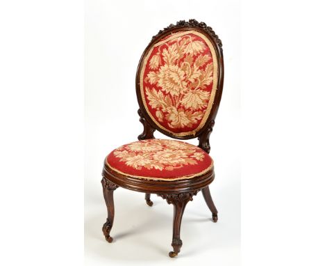 A Victorian carved rosewood nursing chair, the cushioned oval back with floral swag and ribbon cresting, upholstered in flora