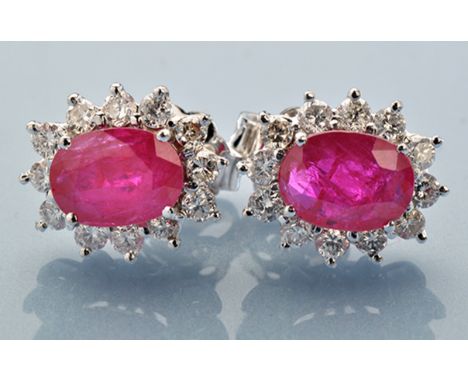 A pair of ruby and diamond cluster earrings, each oval facet cut ruby surrounded by twelve brilliant cut diamonds, in 18ct. w