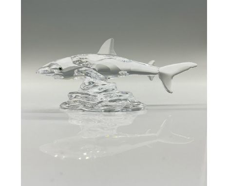 A clear crystal figure of a shark with melon-colored eyes that is swimming over rocks. Includes the original box dimensions: 