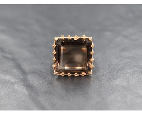 An oversized smoky quartz ring having a square stone in a claw set raised basket mount with engraved shoulders on a gold loop