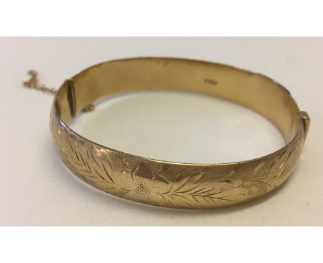 A hallmarked 9ct gold hinged bangle with safety chain. Flower and leaf design engraved to front. Small split in side of bangl