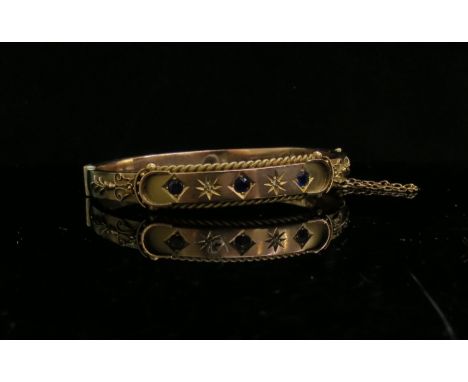 A 9ct gold stiff hinge bangle with foliate scroll detail, set with three blue stones and two old cut diamond chips, dented an