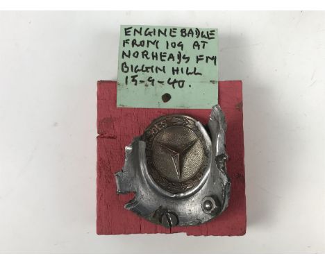 A Mercedes badge crudely mounted on a wooden block with label "Engine badge from 109 at Norheads Fm, Biggin Hill, 19-9-40"