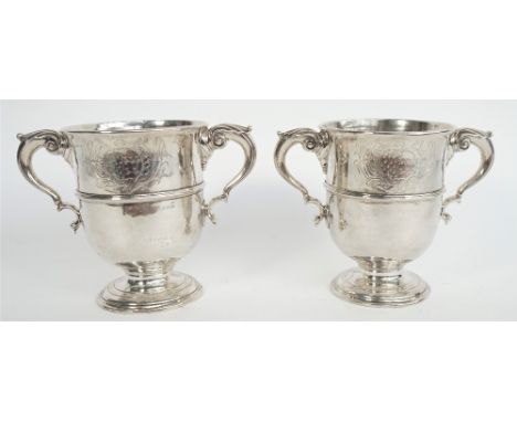 A pair of silver twin handled cups, each body decorated with a central band, with scrolling handles, raised on a circular foo