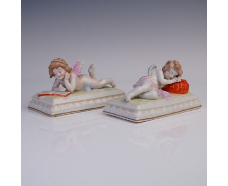 Delightful pair of German porcelain putti figurines, each modeled lying atop a rectangular base with white and gilt trim. One