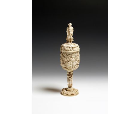 λ A late 19th century German carved ivory cup and cover, with a standing warrior finial, with his trusty hound, above massacr