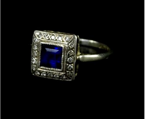 An Art Deco dress ring, with central square cut sapphire, surrounded by tiny diamonds, on a white metal band, marked 18ct pla