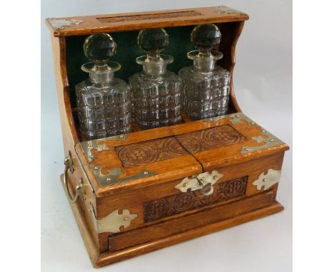 An Edwardian oak three-bottle Tantalus, with silver-plated nickel mounts and side carrying handles, the three square section 