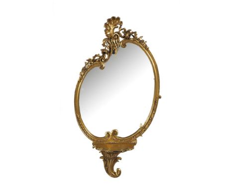 A GILTWOOD SHAPED OVAL WALL MIRROR, the asymmetrical frame with surmounted shell and foliage above a bowfront shelf supported