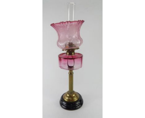A Victorian brass oil lamp with a cranberry glass reservoir, and etched floral cranberry glass shade, raised on a black glass
