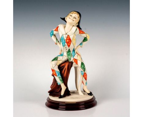 A hand painted figure depicting a young boy sitting on a bench in a harlequin costume. Sculpture is attached on a round woode