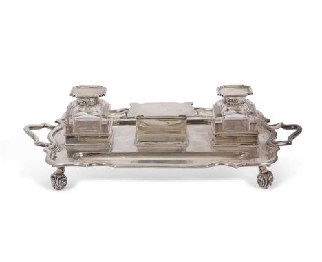 A George V silver two-bottle ink stand of shaped rectangular form, resting on four scroll feet, the square glass silver mount