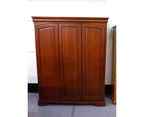 'OLYMPUS' CHERRY WARDROBEwith a moulded top above three panelled doors opening to reveal a fitted interior, standing on brack