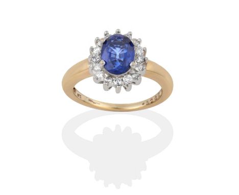 the oval cut sapphire within a border of round brilliant cut diamonds, in white claw settings, to a tapered shoulder plain po