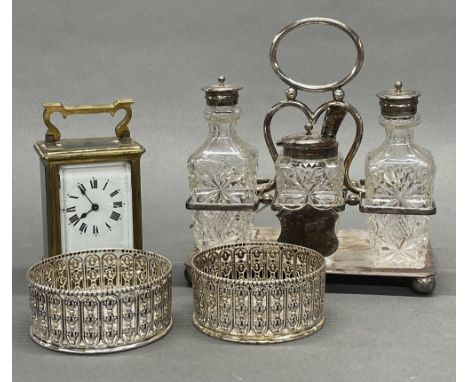 A brass carriage clock with a pair of silver plated wine bottle coasters and a cruet stand.