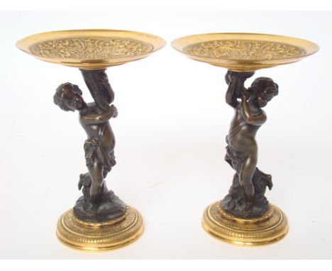 A pair of bronze tazzas, the polished bowls relief cast with chemical figures, on patinated bronze cherub stems and plinth ba