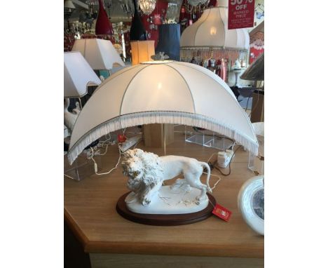 A white ceramic and gold lion table lamp, the lion sits on a large oval wooden plinth and is complete with a cream fringed sh