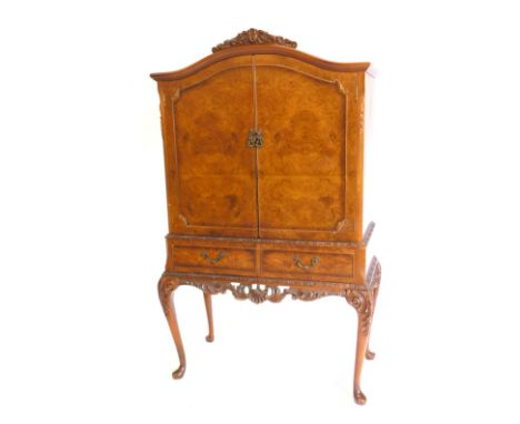 A mid 20thC walnut and burr walnut cocktail cabinet, the arched top with a carved crest, above two doors enclosing a fitted i