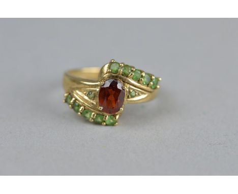 A GARNET AND GREEN STONE 9CT YELLOW GOLD DRESS RING, fancy crossover style, the central oval mixed cut garnet measuring appro