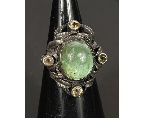 A white metal ring, shank stamped SILVER, with central oval cabochon translucent emerald colour stone surrounded by entwined 