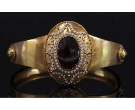 A Victorian gold garnet and enamel hinged bangle, c.1860,with an oval centrepiece. An oval cabochon garnet, rub set to the ce