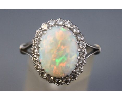 A white metal cluster ring set with an oval cabochon cut opal measuring approximately 14.0mm x 10.5mm and surrounded by twent