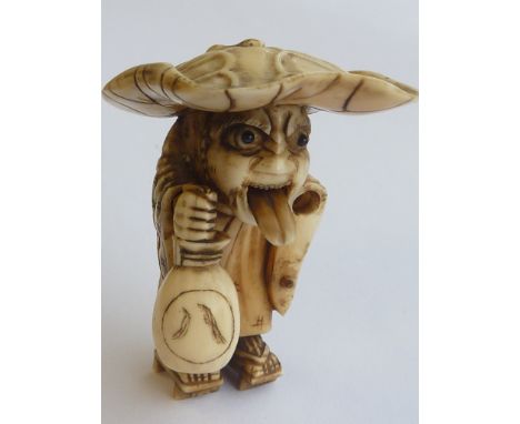 A 19th century carved Japanese ivory Netsuke modelled as a hunched cloaked figure wearing a large lotus leaf shaped hat over 