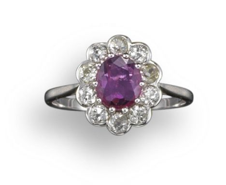 A ruby and diamond cluster ring, the oval-shaped ruby is set within a surround of circular-cut diamonds in white gold, size O