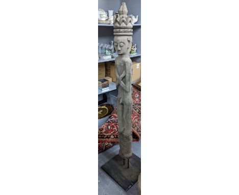 A hardwood carved tribal tall and slender figure depicted standing with arms folded and with a tiered headdress, raised on a 
