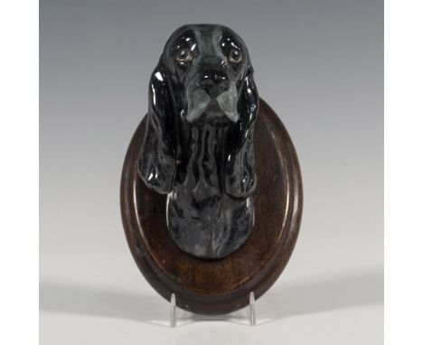 A black glazed figure of a dogs head mounted to an oval wooden panel. Issued: 1930sDimensions: 3.25"W x 5"H x 3.75"DManufactu