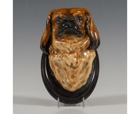 A brown and black glazed figure of a Chinese toy dogs head mounted to an oval wooden panel. Issued: 1930sDimensions: 3.5"W x 