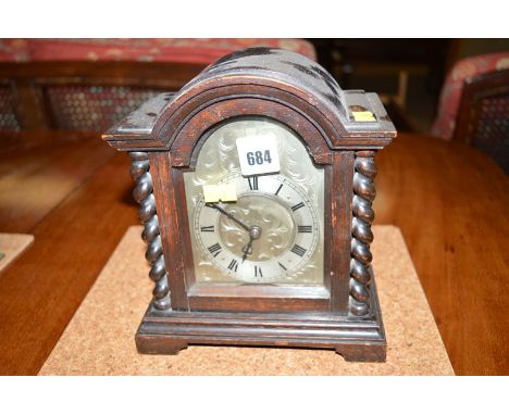 An oak cased mantel clock, the roman dial surrounded by foliate scroll engraving, the arched case above barley-twist columns,