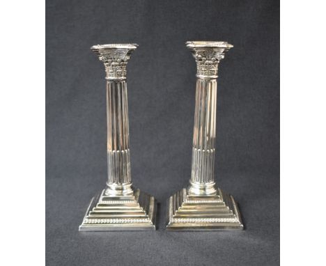 A pair of silver plated Corinthian column candlesticks, having concave square detachable sconces with bead trim, embossed dec