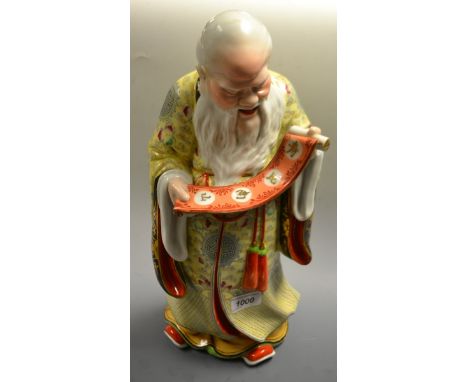 A Chinese porcelain figure, of a scholar, he stands, reading a scroll held in his hands, his long flowing robe decorated with