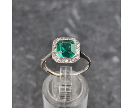 An emerald and diamond cluster ring, the central trap cut emerald of approximately 1.4ct, surrounded by a border of twenty di