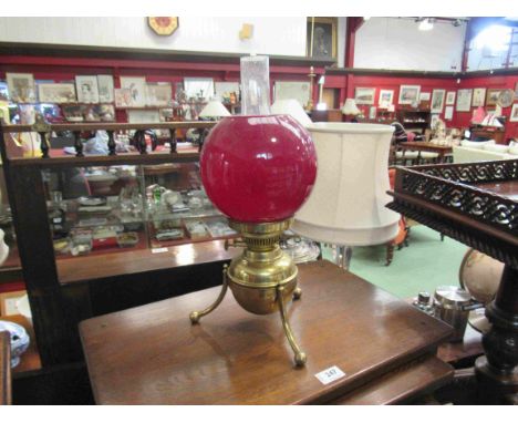 A brass oil lamp on tripod feet with red shade, 43cm tall