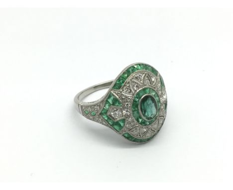 An Art Deco style platinum, emerald and diamond dress ring set with a central oval cut emerald, round cut diamonds and calibr