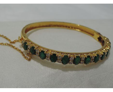 A yellow metal hinged bangle having thirteen oval cut emeralds, total approx 3.5ct  interspersed by pairs of brilliant cut di