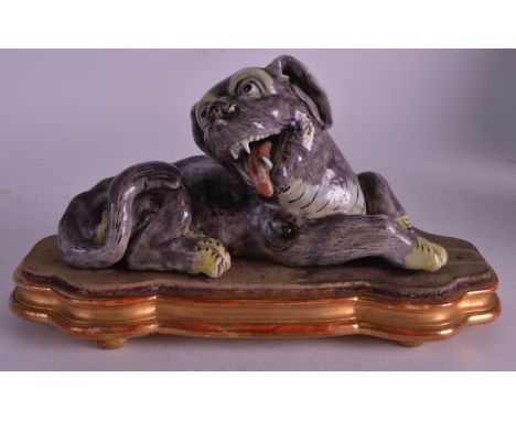 A 17TH/18TH CENTURY CHINESE PORCELAIN FIGURE OF A RECUMBANT HOUND Kangxi, modelled with mouth open, upon a giltwood base. Fig